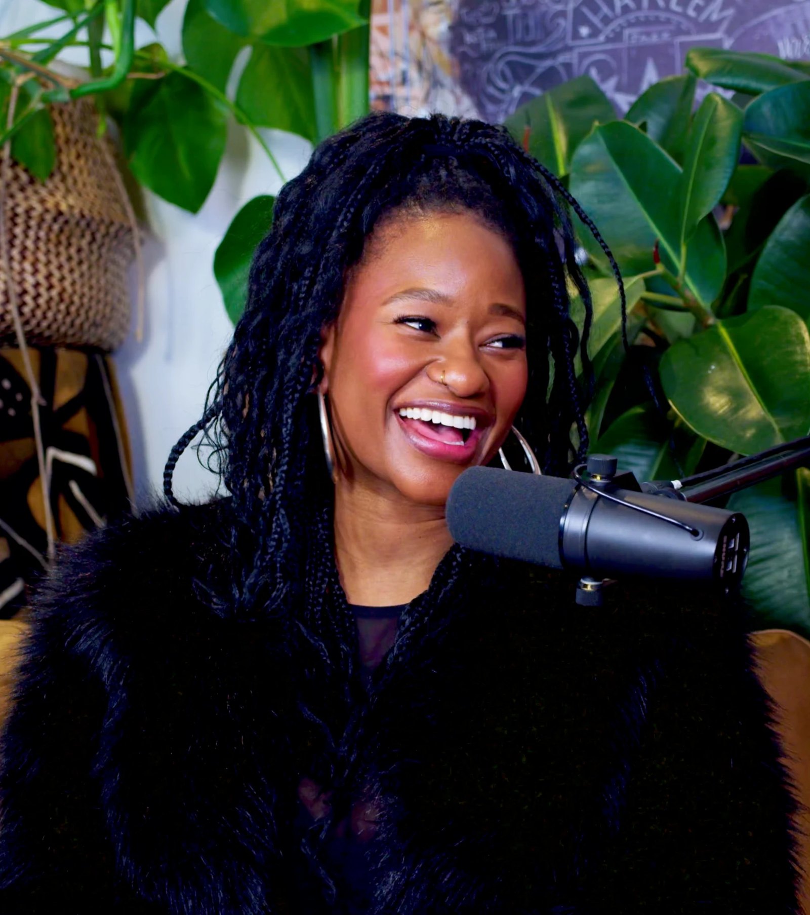 Mati laughing into a studio microphone in a black faux-fur coat, silver hoops and long braids, green plants and a mural behind her